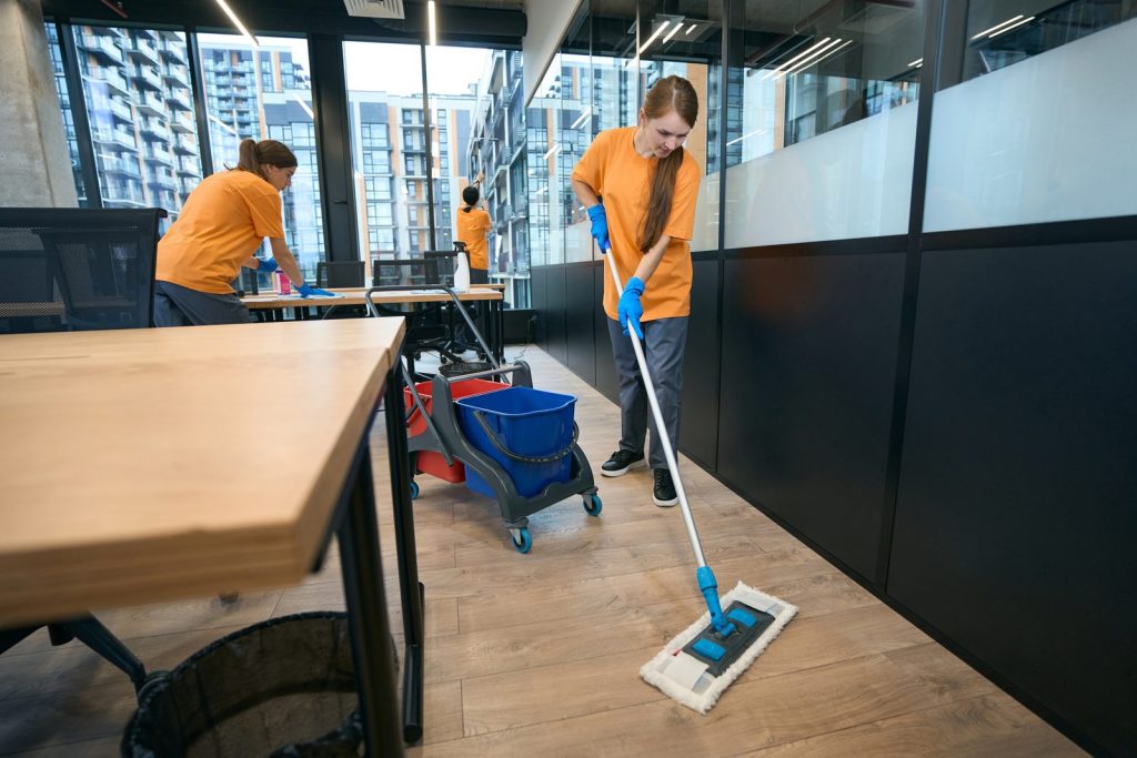 Tres operarias de aseo con camiseta naranja limpian una oficina con ventanales: una trapea el piso, otra limpia un escritorio y otra limpia los vidrios